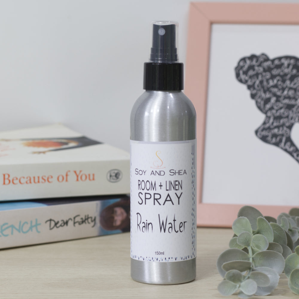 Room and linen spray bottle labeled 'Rain Water' by Soy and Shea on a surface with books and a framed picture in the background.