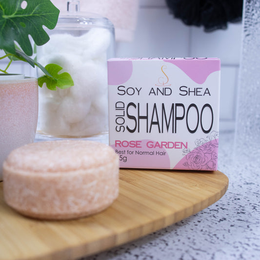 On a wooden tray sits a round light pink Shampoo bar and its cardboard box which is white with an abstract pink design.  Next to it is a potted plant and a jar of cotton balls against a white tile background.