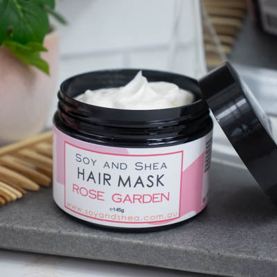 A black jar with the lid resting on its side revealing a white cream inside, sits on a dark grey tile surface against a blurred background of a bathroom counter top. The jar features a white label with pink abstract design with text HAIR MASK ROSE GARDEN.