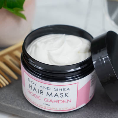 An overhead shot of a black jar with the lid resting on its side revealing a white cream inside, sits on a dark grey tile surface against a blurred background of a bathroom counter top. The jar features a white label with pink abstract design with text HAIR MASK ROSE GARDEN.
