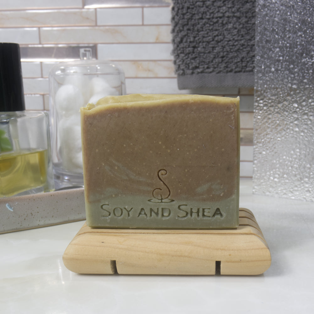 A rectangular bar of soap stands on a wooden soap dish in front of some bathroom accessories including a bottle of perfume. The soap has a dark olive green layer toped with a brown-green colour on top. 