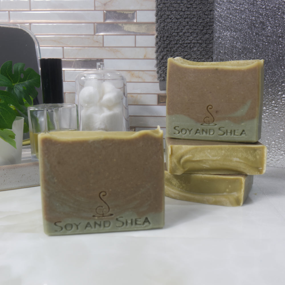 A selection of soaps are displayed on a bathroom countertop along with a bottle of perfume and jar of cotton balls. One soap stands upright on the bench, while behind and to the left, two bars lay flat stacked upon each other revealing an bright olive green top with slght wavy texture.  Another soap stands upright on these.  They feature a design of a small layer of  forest green across the base of the soap with the top being a dark olive green.