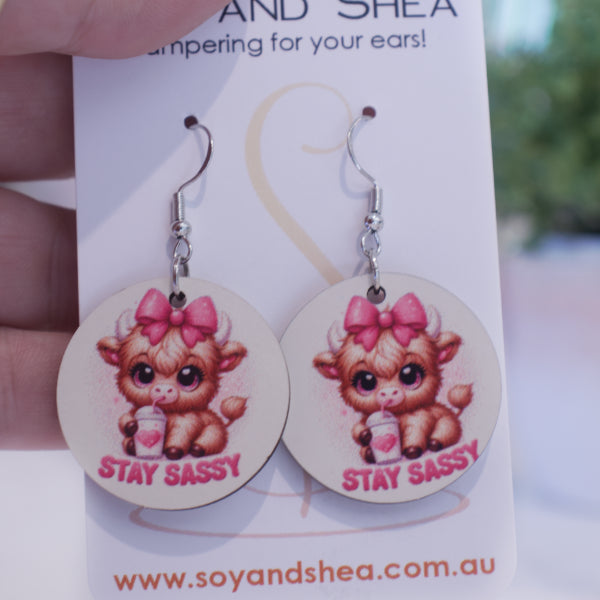 A hand holds a white card branded with SOY AND SHEA with a pair of earrings hanging on hooks.  The earrings are round with an illustration of a baby highland cow with a pink bow on its head drinking from a white cup, with heart and straw.  The words STAY SASSY are curved along the bottom.