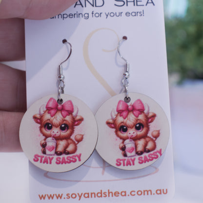 A hand holds a white card branded with SOY AND SHEA with a pair of earrings hanging on hooks.  The earrings are round with an illustration of a baby highland cow with a pink bow on its head drinking from a white cup, with heart and straw.  The words STAY SASSY are curved along the bottom.