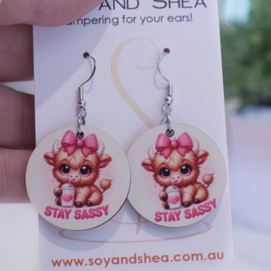 A hand holds a white card branded with SOY AND SHEA with a pair of earrings hanging on hooks.  The earrings are round with an illustration of a baby highland cow with a pink bow on its head drinking from a white cup, with heart and straw.  The words STAY SASSY are curved along the bottom.