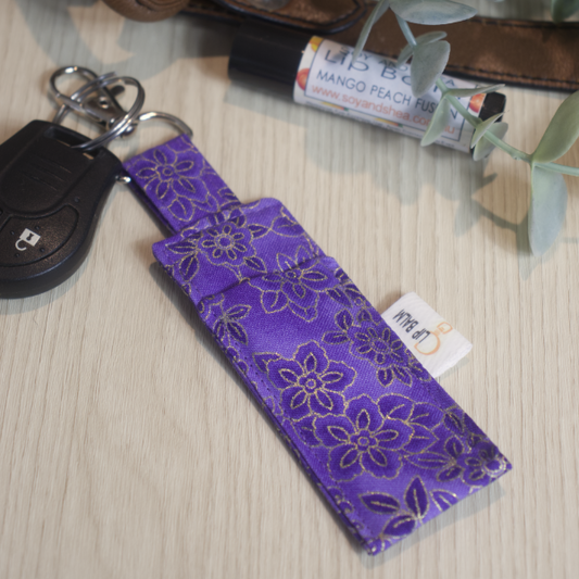 A clip balm holder lies on a wooden surface attached to a car key with a "lobster claw" clip.  To the side is a tube of lip balm surrounded by greenery and a leather strap from a bag.  The Clip Balm has a royal purple fabric with a slightly darker floral design outlined in gold