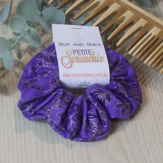 A colorful scrunchie sits on wooden surface resting against a wooden comb with greenery in the background.  A cardboard tag reads SOY AND SHEA PETITE SCRUNCHIE.    The scrunchie is a royal purple fabric with a floral design a shade darker and outlined with gold