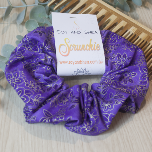 A colorful scrunchie sits on wooden surface resting against a wooden comb with greenery in the background.  A cardboard tag reads SOY AND SHEA SCRUNCHIE.    The scrunchi is a royal puple colour featuring a slightly darker floral design across the fabric outlined in gold