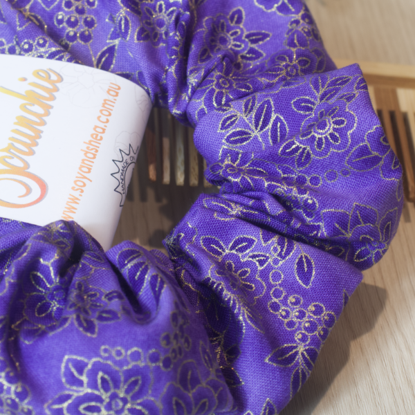A close up of the Royal Bloom Scrunchie showing the desing in detail.  The scrunchie has a royal purple background with a slightly darker floral pattern outlined in gold
