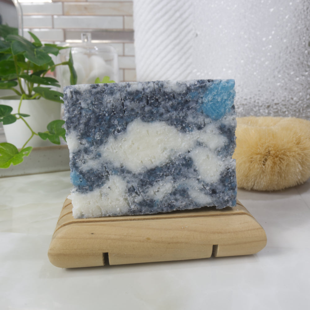 A bar of soap stands on a wooden soap dish on a bathroom counter top.  The soap has a mottled appearance in colours of grey, white and teal