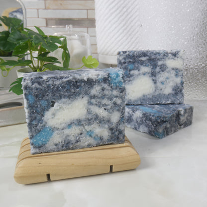 A selection of soap bars are displayed on a bathroom counter next toa plant and jar of cotton balls.   In the foregrund is a soap standing on a wooden soap dish and behind to the right are two more soaps. One lying flat and another standing upon it.  The soaps have a mottled appearance in colours of White, grey and teal.