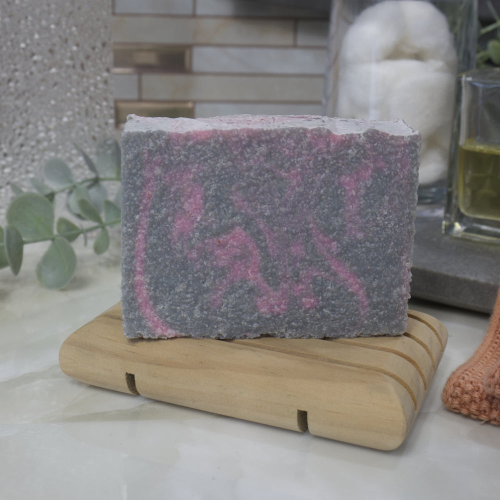 Gray and pink soap bar with textured surface sits on a wooden stand with a blurred background