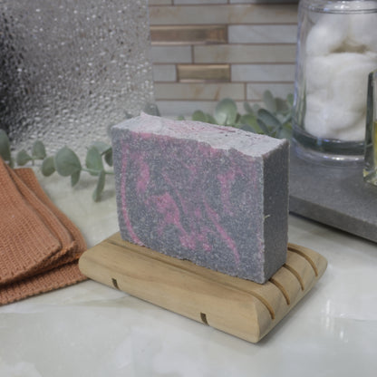 Bar of soap on a wooden stand with a textured background. The soap is grey with pink swirls throughout