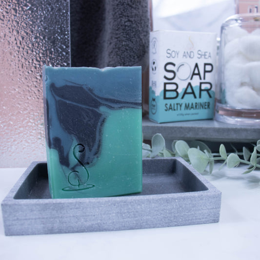 A tall rectangular bar of soap sits on a grey soap dish. The soap has three layers of colour; a sea green base, black centre and teal at the top, which have been swirled together to look like a rolling wave.  Behind the soap is a shower screen and towel along with a box the soap was presented in. The box has a white top with sea green bubble border.