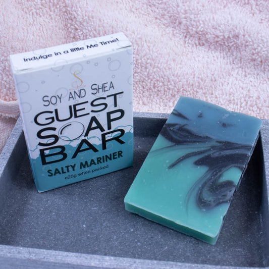 A grey stone soap dish sits next to a pink towel.  On the dish stands a white box with a border of ocean green bubble foam across the bottom and text GUEST SOAP BAR. Next to it lies a small piece of soap  that has a bottom layer of ocean green with black and dark green swirling into it from the top.