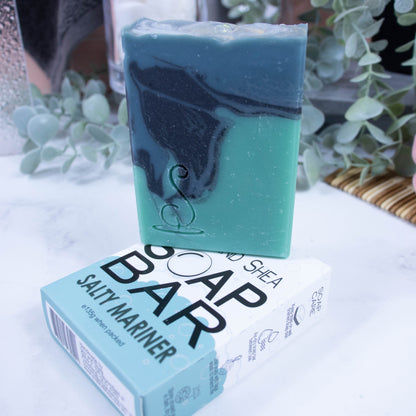 A tall rectangular soap bar is sitting on top of it's box.  The soap has three layers of colour; a sea green base, black centre and teal at the top, which have been swirled together to look like a rolling wave.  The soap box is laying flat and is white and with a sea green bubble border.