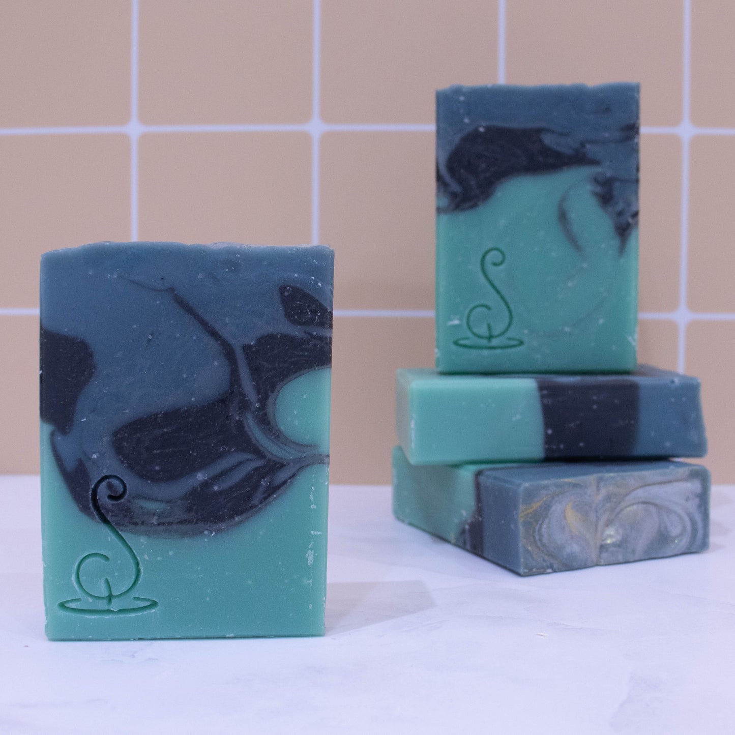Four rectangular bars of soap sit on a marble bench each showing various design patterns from the same batch of soap. Two soaps lay flat but stacked upon each other showing a flat top.  A third soap stands on these while a fourth is presented in front of the stack.  The soaps have three layers of colour; a sea green base, black centre and teal at the top, which have been swirled together to look like a rolling wave.  All soaps have a similar pattern but all have slight differences