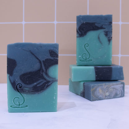 Four rectangular bars of soap sit on a marble bench each showing various design patterns from the same batch of soap. Two soaps lay flat but stacked upon each other showing a flat top.  A third soap stands on these while a fourth is presented in front of the stack.  The soaps have three layers of colour; a sea green base, black centre and teal at the top, which have been swirled together to look like a rolling wave.  All soaps have a similar pattern but all have slight differences