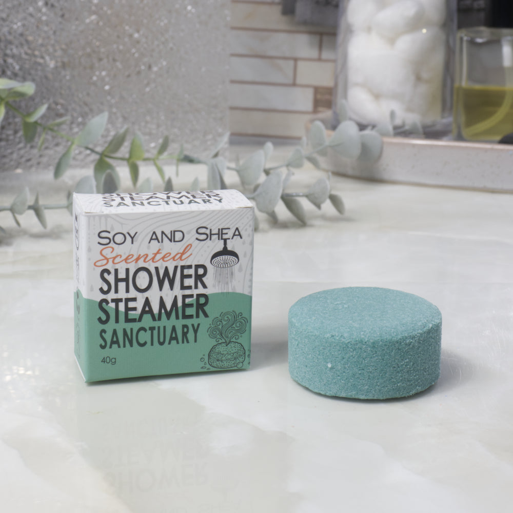 A boxed scented shower steamer and the steamer itself on a bathroom counter. The box reads 'Soy and Shea Scented Shower Steamer Sanctuary', and the round dark green steamer sits beside it.