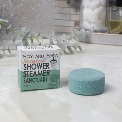 A boxed scented shower steamer and the steamer itself on a bathroom counter. The box reads 'Soy and Shea Scented Shower Steamer Sanctuary', and the round dark green steamer sits beside it.