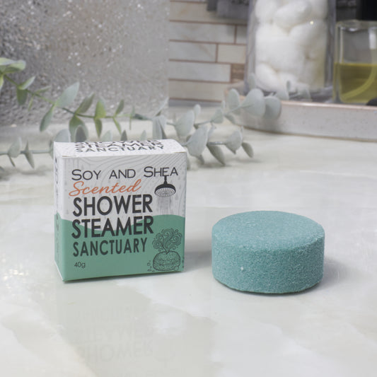 A boxed scented shower steamer and the steamer itself on a bathroom counter. The box reads 'Soy and Shea Scented Shower Steamer Sanctuary', and the round dark green steamer sits beside it.