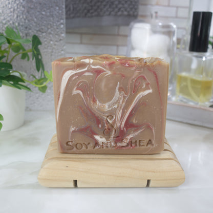 A bar of soap stands on a wooden soap dish with a blurred bathroom background.  The soap is a light brown with swirls of dark brown, dark red and white swirls.