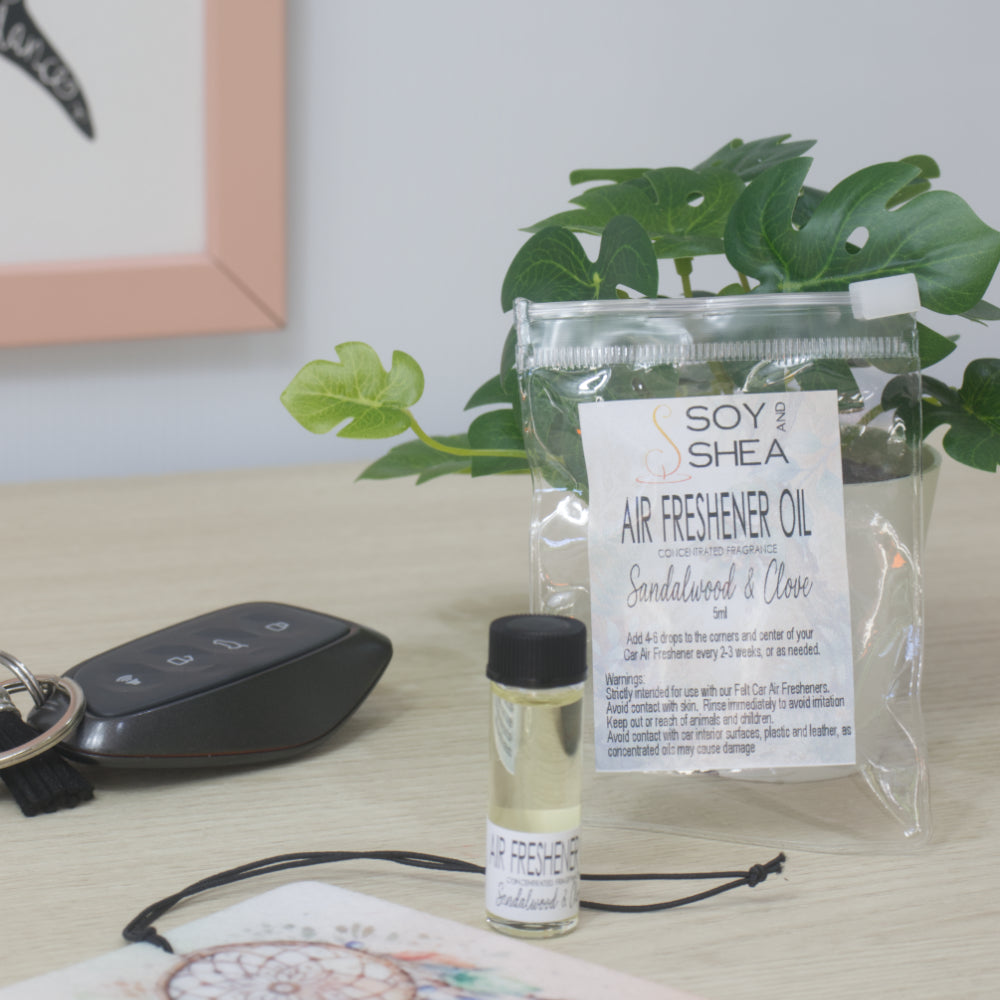A small clear bottle with black cap of Soy's Shea Air Freshener Oil next to a clear resealable pouch, displayed on a light wood tabletop with a car key, a felt air freshener, and a potted plant in the background.  