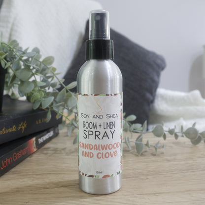 An aluminium bottle with black spray cap stands on a wooden surface next toa pile of books with a plant sitting on top of them.  A chair with white blanket and grey cushion is blurred in the background.   The bottle features a decorative product label identifying it as Room + Linen Spray, scented in Sandalwood and Clove