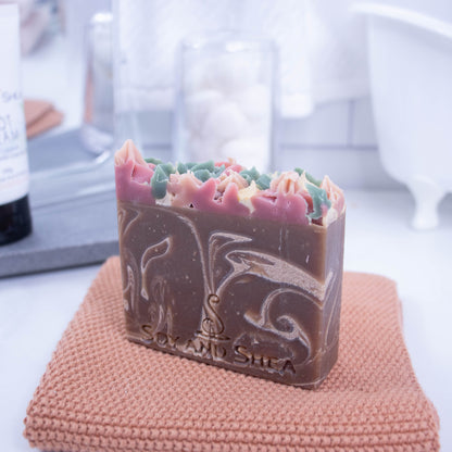  A bar of soap stands upgright on a rusty orange face cloth with a bright blurred background of a bathroom countertop.  The soap has a base colour of chocolate brown with cream swirls and features a piped soap top of pink, cream and green.