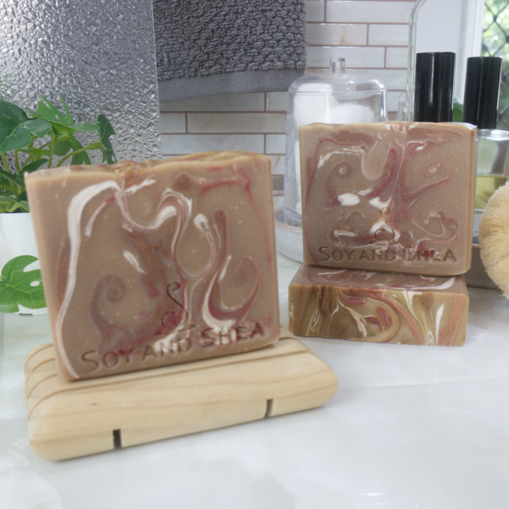 A selection of three soaps displayed on a bathroom bendch along with a bottle of perfume, plant and jar of cotton balls.  One soap sits on a wooden rack to the front left of the image, another two are displayed behind with one laying flat showing the top of the soap with a third standing on top.  The soaps are a light brown with varying swirls of dark red, dark brown and white.  The tops of the soap are flat with colour swirls.