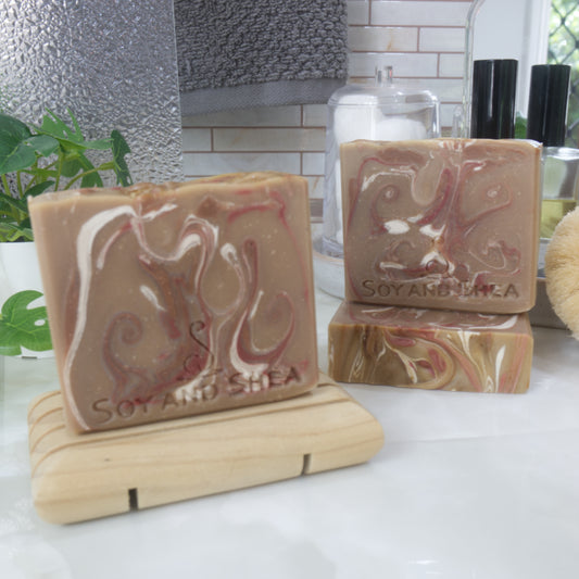 A selection of three soaps displayed on a bathroom bendch along with a bottle of perfume, plant and jar of cotton balls.  One soap sits on a wooden rack to the front left of the image, another two are displayed behind with one laying flat showing the top of the soap with a third standing on top.  The soaps are a light brown with varying swirls of dark red, dark brown and white.  The tops of the soap are flat with colour swirls.