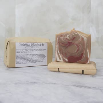 A brown soap bar with red and white swirls sits on a wooden stand with a brown paper package behind labeled 'Sandalwood and Clove Soap Bar' on a marble background