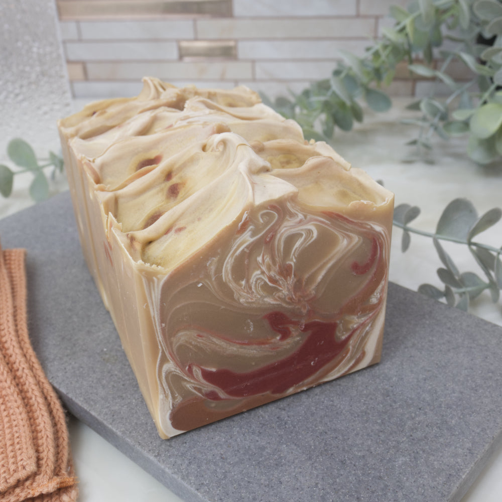Swirled soap bar on a gray surface with greenery in the background.  The front soap is light brown in colour with decorative swirls in red, brown and white.