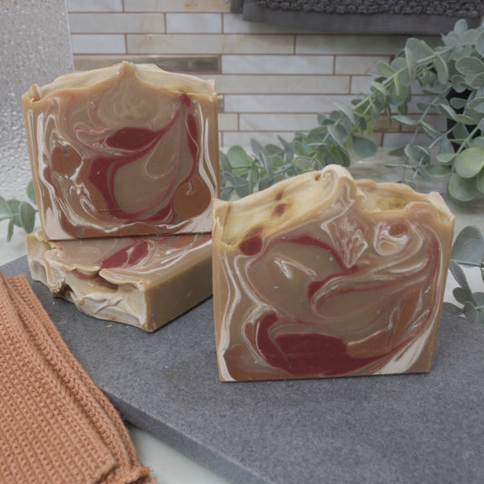 Three soap bars sit on a stone surface surrounded by greenery and towels with a tiled background.  One soap lays flat on the stone surface showing the textured wavy top.  Another stands on top of this soap showing a swirl design of a tan base with red, brown and white swirls. In front is another soap with the same colours but different pattern