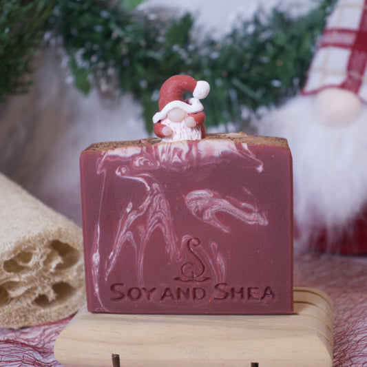 The Santa's Grotto Soap Bar (Christmas Edition) features a festive marbled red and white design, crowned with a small Santa figurine. Made by Soy and Shea, this Christmas aroma soap is handmade with plant-based oils and elegantly arranged on a wooden dish surrounded by greenery and red holiday accents.