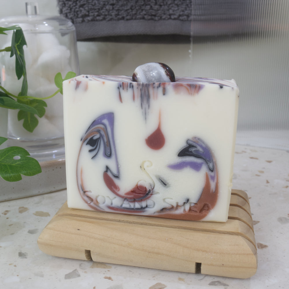 A bar of soap stands on a wooden soap dish on a bathroom countertop.  The soap is white with drops of colour in browns, black and purple.  On the top centre, a sardonxy stone is embedded.