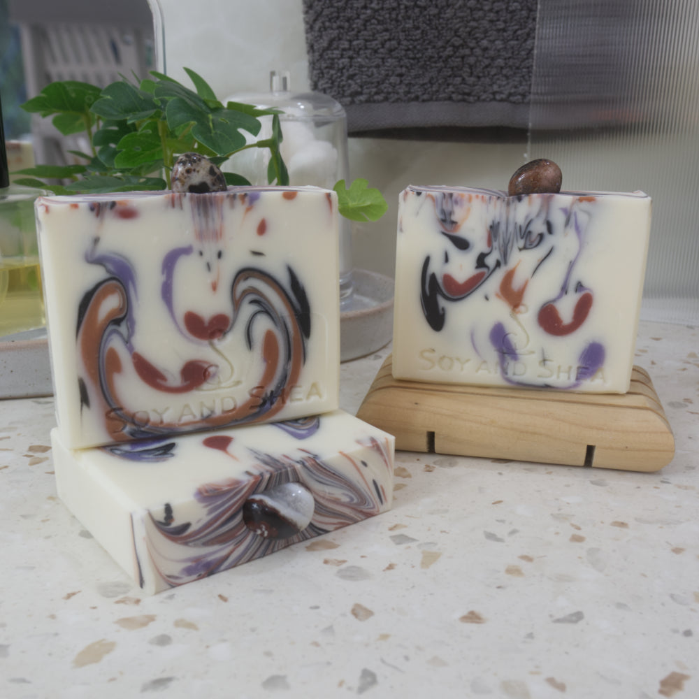 A selection of soaps are displayed on a bathroom countertop with a plant.  To the front left, a soap lays flat on the bench showing a colourful soap top with a piece of Sardonyx embedded in it. On this stands a soap and to the right back, another soap stands on a wooden soap rack.  The soaps all have a white background with various drops of colour in browns, black and purple matching th colours of the stones on each soap.