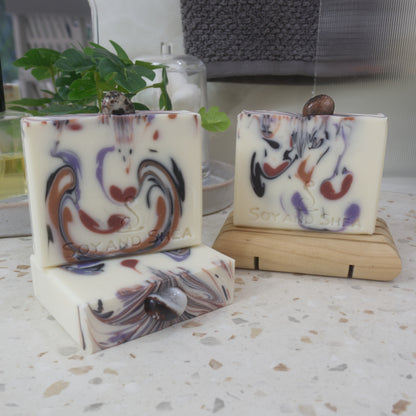 A selection of soaps are displayed on a bathroom countertop with a plant.  To the front left, a soap lays flat on the bench showing a colourful soap top with a piece of Sardonyx embedded in it. On this stands a soap and to the right back, another soap stands on a wooden soap rack.  The soaps all have a white background with various drops of colour in browns, black and purple matching th colours of the stones on each soap.