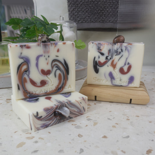 A selection of soaps are displayed on a bathroom countertop with a plant.  To the front left, a soap lays flat on the bench showing a colourful soap top with a piece of Sardonyx embedded in it. On this stands a soap and to the right back, another soap stands on a wooden soap rack.  The soaps all have a white background with various drops of colour in browns, black and purple matching th colours of the stones on each soap.