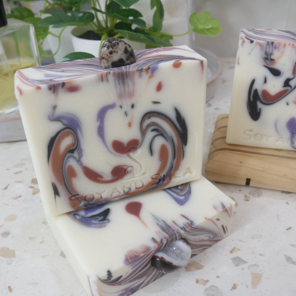 A close up image of two soaps that have a white base colour with colourful drops in shades of brown, black and purple.  Each soap has a sardonyx stone embedded in them