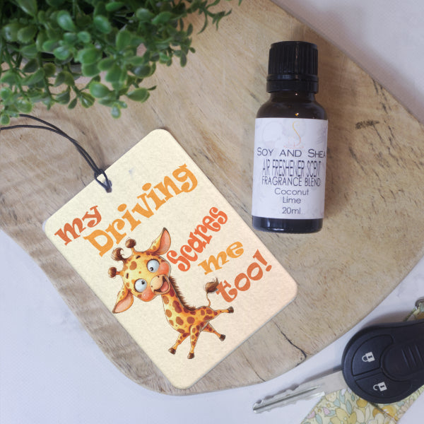 A car air freshener featuring crazy looking cartoon giraffe is surrounded by the words  MY DRIVING SCARES ME TOO! in shades of orange is placed on a wooden surface.  Next to is a glass bottle of Air Freshener Scent  
