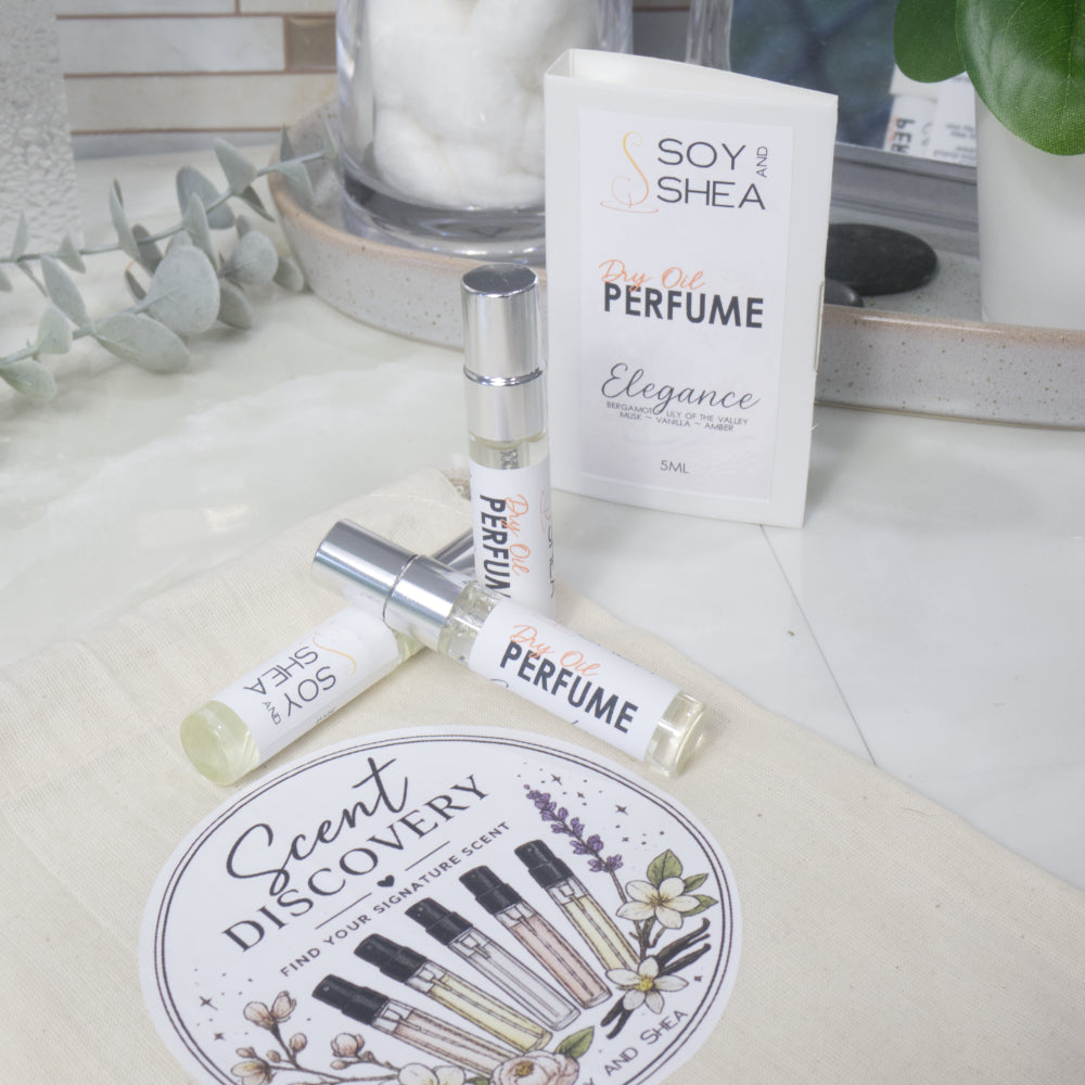 Three small clear Dry Oil Perfume atomizer bottles with silver caps are arranged on a calico bag labelled as Scent Discovery.  Behind is a card labelled as Dry Oil Perfume Elegance surround by bathroom accessories