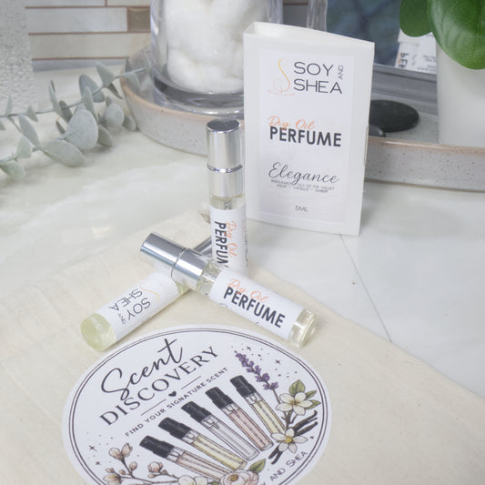 Three small clear Dry Oil Perfume atomizer bottles with silver caps are arranged on a calico bag labelled as Scent Discovery.  Behind is a card labelled as Dry Oil Perfume Elegance surround by bathroom accessories
