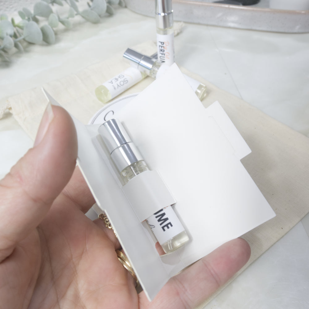 A hand holds a small perfume sample vial in a white paper sleeve, with additional perfume bottles and a neutral fabric background.