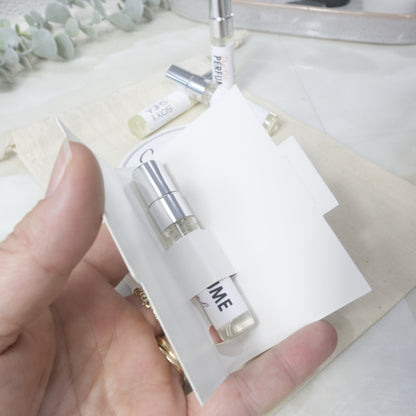A hand holds a small perfume sample vial in a white paper sleeve, with additional perfume bottles and a neutral fabric background.