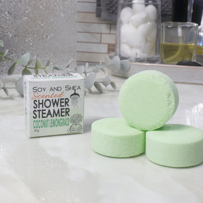 A box of Soy and Shea Scented Shower Steamer in Coconut Lemongrass next to three round, pale green shower steamers arranged on a white marble-like surface.