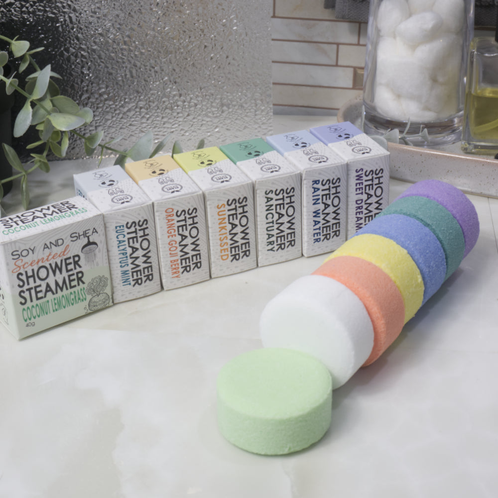 A set of round shower steamer discs in pastel rainbow colors arranged in front of their branded boxes on a bathroom counter. The boxes are labeled with different scents including Coconut Lemongrass, Sunkissed, and Sweet Dream.