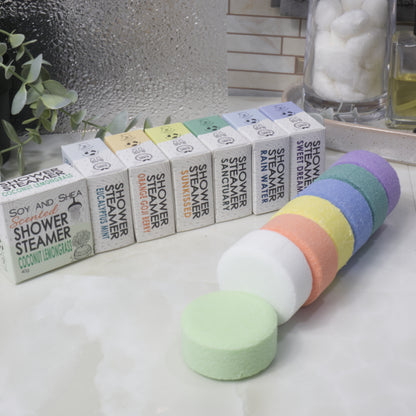 A set of round shower steamer discs in pastel rainbow colors arranged in front of their branded boxes on a bathroom counter. The boxes are labeled with different scents including Coconut Lemongrass, Sunkissed, and Sweet Dream.
