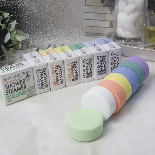 A set of round shower steamer discs in pastel rainbow colors arranged in front of their branded boxes on a bathroom counter. The boxes are labeled with different scents including Coconut Lemongrass, Sunkissed, and Sweet Dream.