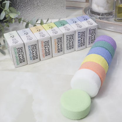 Seven small rectangular boxes of shower steamers are lined up behind a stack of round shower steamer discs in pastel colors, with one pale green pad placed in front on a light marble-like surface.